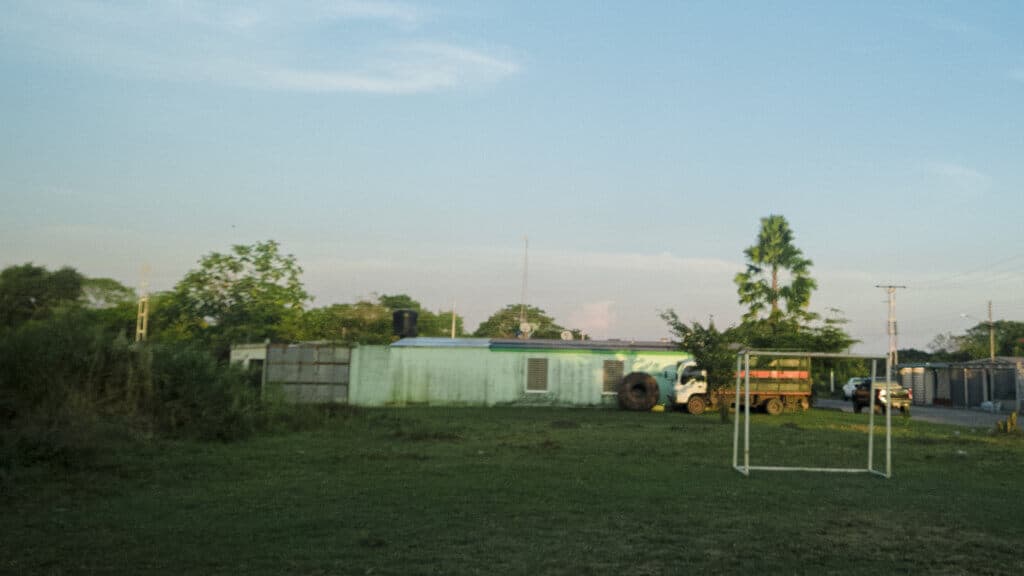 Hierba y árboles en un campo con construcciones modestas y un pequeño arco deportivo en un entorno rural.