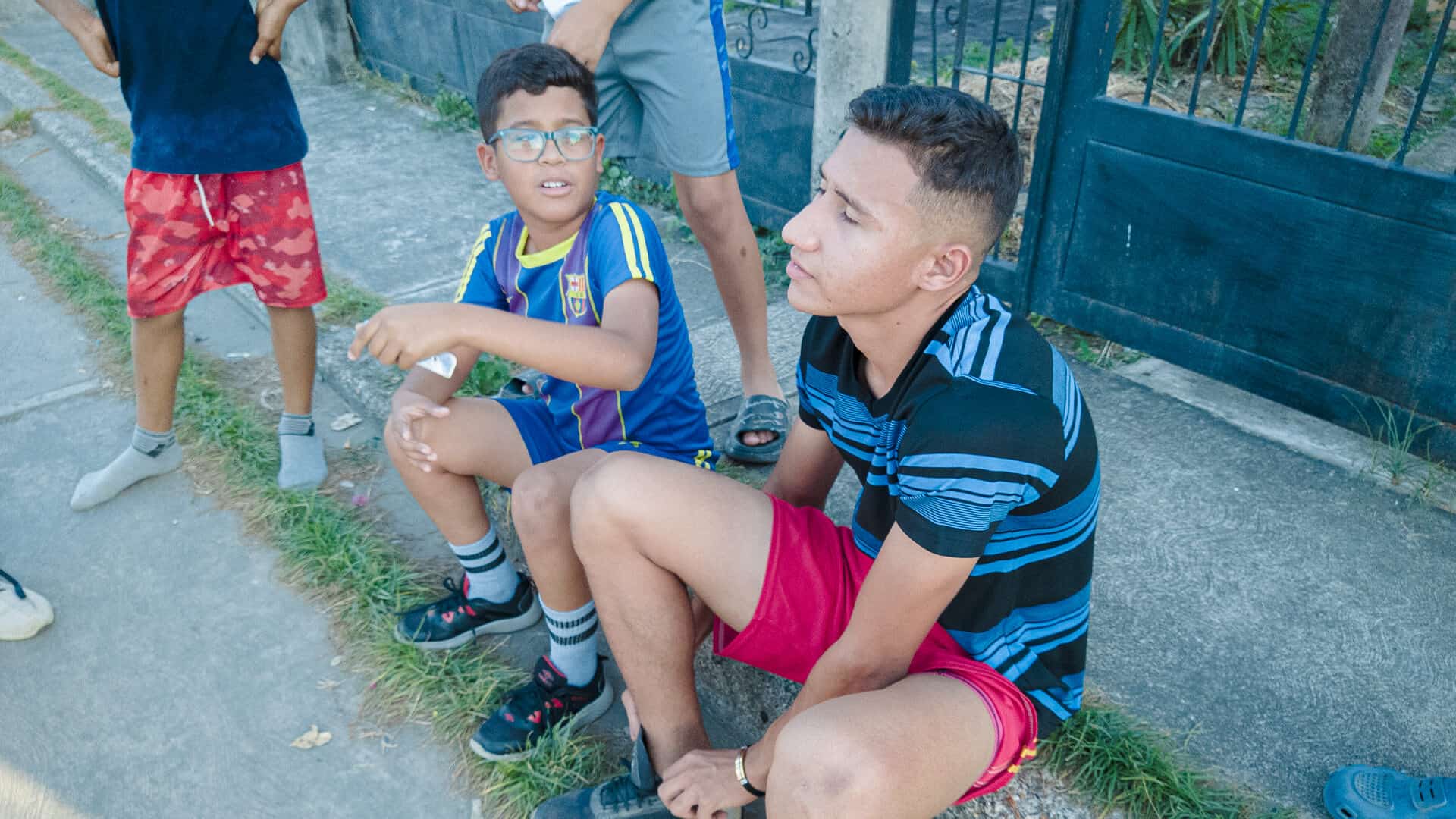Joven sentado en la acera con niños y adultos en el fondo, en una escena cotidiana en Apure, Venezuela.
