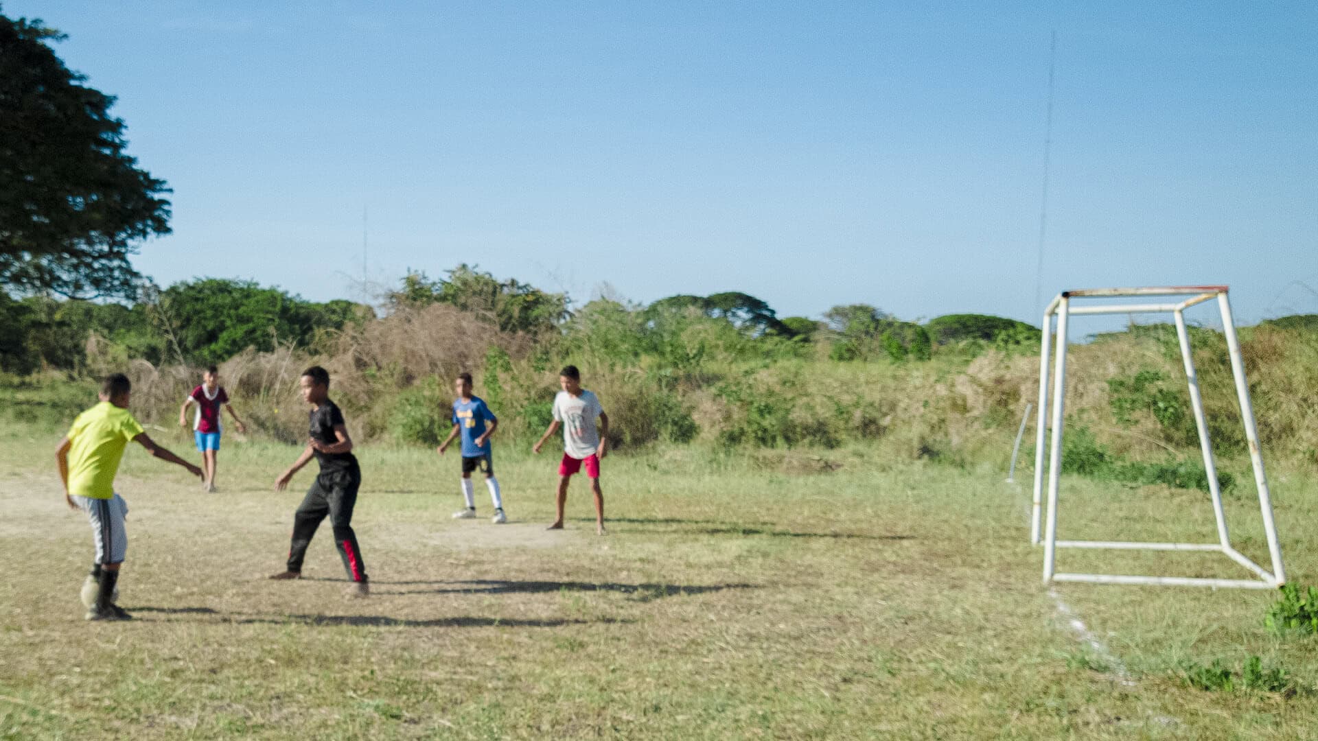 Niños jugando fútbol en campo rústico en Apure con fondo de vegetación y cielo despejado.